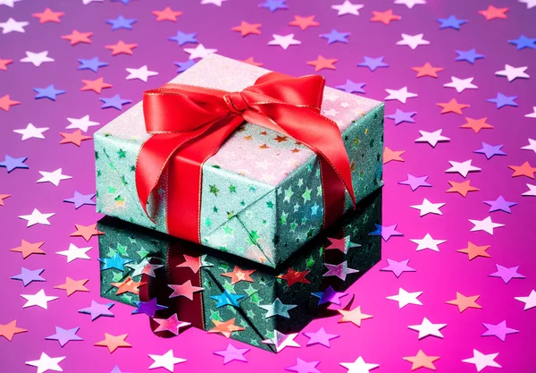 Single green gift box with red ribbon bow on purple reflective surface with glitter stars. Gift box wrapped in green shine paper with red ribbon close up
