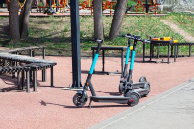 Elektrikli scooterlar parka park edilmiş, kiralık, arka planda egzersiz ekipmanları var.