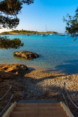 Access stairs to the Xinxell cove, among pine trees, moored sailboats and the Sa Torre island with its tower