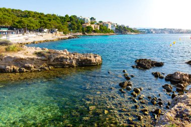 Rocky coast of the Cala Comtesa cove from the Punta Des Bufador isthmus, buildings among pine trees in the Ses Illetes cove. Cala Major cove in the background