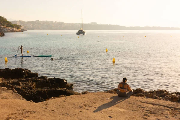 Ses Illetes, Mayorka, Balear Adaları, İspanya. 22 Temmuz 2022 - Katamaran ve Cala Kontes Koyu 'nun kayalık kıyılarında sörf yapan insanlar, gün doğumunda yoga yapan bir kadınla birlikte
