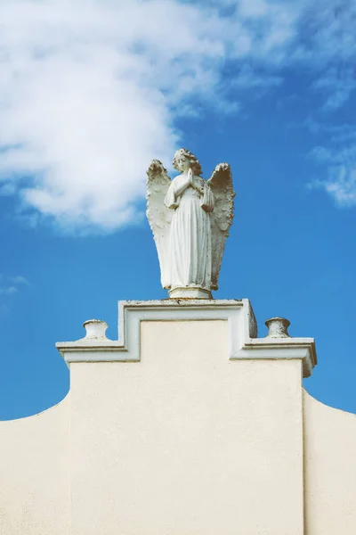 Female sculpture of an praying angel on a white wall with copy space, with blue sky with clouds in the background
