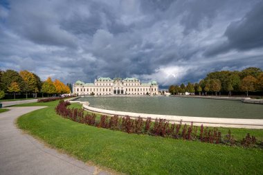 Avusturya, Viyana 'daki Belvedere Sarayı' nın bir kenar manzarası, sonbahar ağaçları tarafından çerçevelenmiş ve önündeki sakin su, sarayın mimarisinin ihtişamını ve doğal güzelliği vurguluyor.