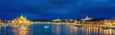 Macaristan 'ın Budapeşte kentinin panoramik manzarası, aydınlanmış Macar Parlamento Binası ve Tuna Nehri' nin karşısındaki şehrin manzarası, birkaç dağınık bulutla dolu, sakin bir gece gökyüzünün altında.