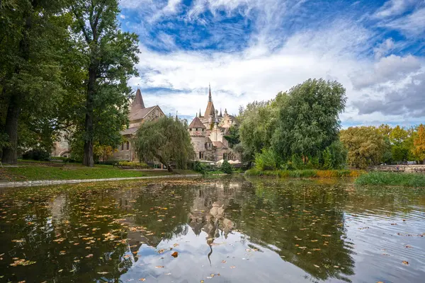 Vajdahunyad Şatosu 'nun önünde sonbahar yapraklarıyla süslenmiş, bulutlu bir gökyüzüne bakan, Budapeşte, Macaristan' ın dingin güzelliğini yansıtan küçük bir göl.
