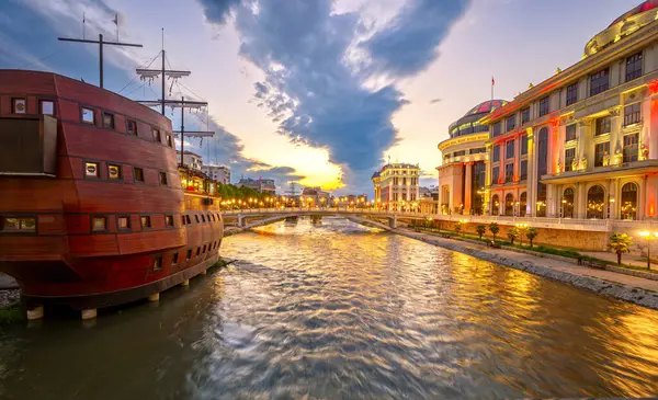 Kuzey Makedonya 'nın başkenti Üsküp' ün güzel bahar şehri, gün batımında gökyüzünde canlı renkler ve Vardar Nehri 'nin sularında yansımalarla.