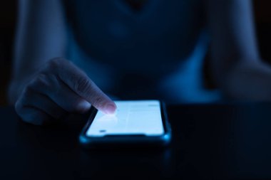 Geceleri cep telefonuyla çalışan bir kadın, karanlık geçmişi olan bir akıllı telefona dokunan bir parmak.