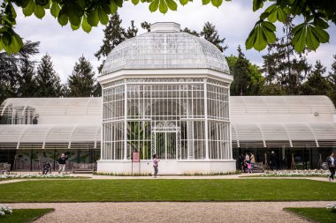 Viktorya tarzı beyaz bir sera. Etrafta çok az insan dolaşıyor. Paris 'te bir parkta çekildi Bulutlu bir bahar gününde.