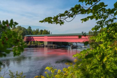 A framed view on a red wooden covered bridge over a large river with the boreal forest around it, Laurentians, Quebec, Canada. Taken on a sunny summer afternoon with no people.