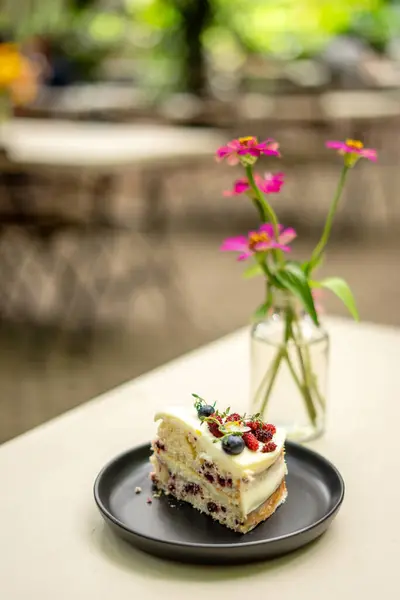 A slice of Vanilla Mixed Berry Cake adorned with mulberries on top, revealing the layers of mixed berries and cream, as well as the soft, tender cake beneath, with flower vase and blure background.