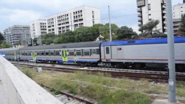  Mavi ve gri yolcu treni, arka planda binalar, ağaçlar ve üstteki elektrik hatlarıyla şehir merkezindeki tren raylarının üzerinde duruyor..