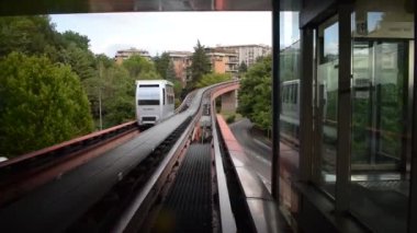 Beyaz minimetro treni, bulutlu bir gökyüzü altında yeşil ağaçlar ve deniz mavisi apartmanlar arasında yükseltilmiş bir yolda ilerliyor..