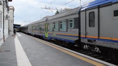 Hareket halindeki mavi ve turuncu çizgili yolcu treninin elektrik hatlarının altındaki istasyon platformunda görüntüsü..