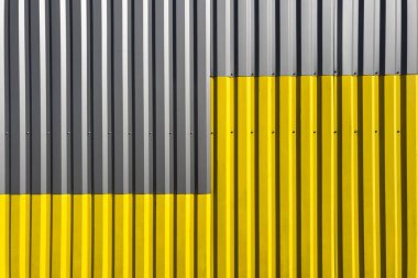 Endüstriyel kıvrımlı duvar, dikey çizgiler, yarısı gri ve yarısı parlak sarıya boyanmış, cesur bir geometrik karşıtlık oluşturuyor.