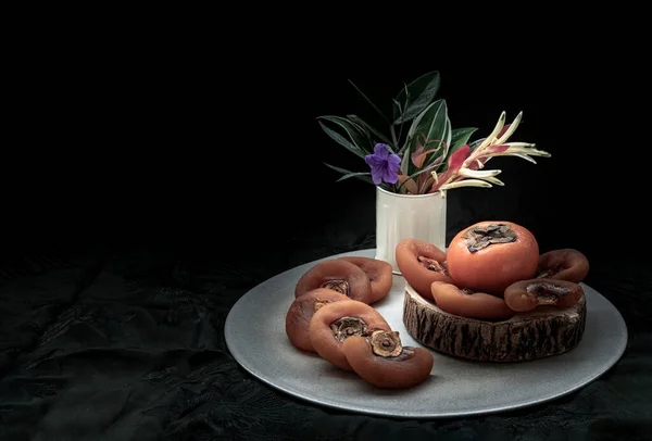 stock image One fresh ripe persimmon fruit and Dried persimmons wity Colorful flower vase on round ceramic tray. Space for text, Selective Focus.