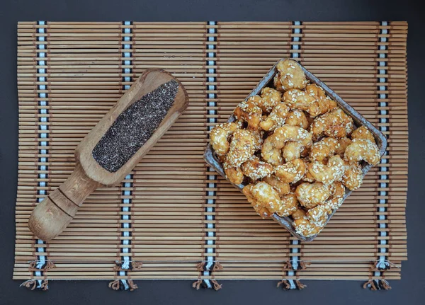 Sweet Cashew Nuts coated with white sesame seeds in the ceramic bowl served with black sesame on bamboo mat. Healthy snack, Top view, Space for text, Selective Focus.