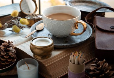Morning refreshing with Hot coffee in ceramic cup and brass coffee spoon served with Fresh milk on worktable with old book, brass compass, scented candle and dried flowers. Selective focus.