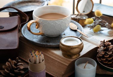 Morning refreshing with Hot coffee in ceramic cup and brass coffee spoon served with Fresh milk on worktable with old book, brass compass, scented candle and dried flowers. Selective focus.