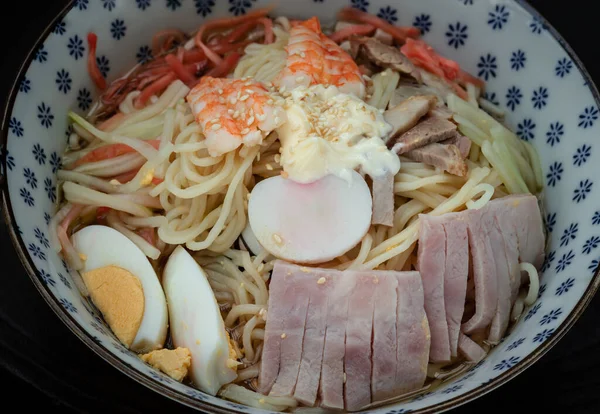 Cold noodles (Remen) with roast pork sliced, shrimp, ham, boiled egg and lemon sauce in blue ceramic bowl served with spoon and chopsticks on black background. Japanese style noodles, Healthy life style eating and Asian food concept. Space for text,