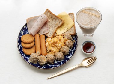 Ev yapımı kahvaltıda ekmek, kızarmış sosis, jambon, domuz shumais, omlet, peynir, çıtır zencefilli kurabiye ve beyaz arkaplanlı bir fincan sağlıklı protein karışımı sunulur. Sağlıklı gıda konsepti, metin için boşluk, Seçici Odaklanma.