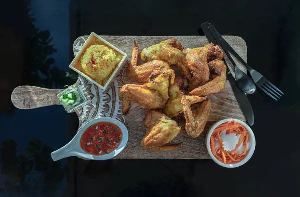 Delicious Fried chicken wings served with Homemade yellow jasmine rice (Chicken biryani), Pickled carrot and Radish, Cucumber sticks and Sweet chili sauce on a wooden cutting board. Halal food, Top view, Copy space, Selective focus.