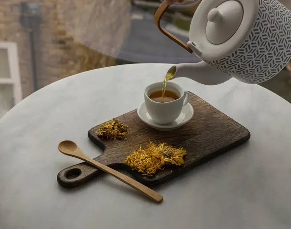 Pouring Chinese chrysanthemum flower tea from Ceramic teapot into a white teacup on wooden cutting board. Dried chrysanthemum flower, Healthy beverage for drink, Space for text, Selective focus.