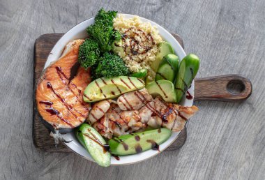 Kahvaltıda somon bifteği, tavuk bifteği, kuskus, kesilmiş Avocado, Brokoli ve seramik tabağa serpiştirilmiş salatalık sirkesi yer alıyor. Organik ve sağlıklı yiyecekler. Üst görünüm, metin için boşluk, Seçici odak.