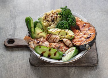 Kahvaltıda somon bifteği, tavuk bifteği, kuskus, kesilmiş Avocado, Brokoli ve seramik tabağa serpiştirilmiş salatalık sirkesi yer alıyor. Organik ve sağlıklı yiyecekler. Yukarıdan belirsiz görünüm, boşluğu kopyala, Seçici odak.