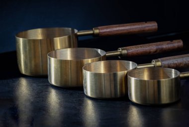 Koyu arkaplanda Asılı Delik Tasarımı ile ahşap saplı Pirinç Ölçme Bardakları. Kuru ve Sıvı İçindekileri Ölçmek için Tahta & Pirinç Ölçme Kadehleri ve Kaşıklar, Mutfak aracı, metin için alan, Seçici Odaklanma.