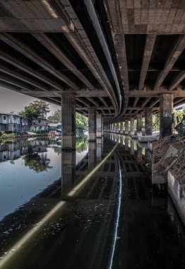 Bangkok, Thailand - Apr 20, 2024 - Perspective view of concrete pillars under Khlong Toei Expressway with calm surface water reflection and houses are old along the Khlong Phra Khanong. Space for text, Selective focus. clipart