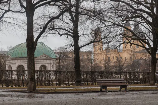 stock image Munich, Germany - Dec 22, 2023 - Bench made wood and iron stand in the garden of the Munich Residenz with The Diana Temple (Dianatempel) and St.Cajetan and Adelaide in the background. Space for text, Selective focus.