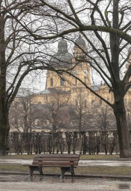 Munich, Germany - Dec 22, 2023 - Bench made wood and iron stand in the garden of the Munich Residenz with St.Cajetan and Adelaide in the background. Copy space, Selective focus. clipart