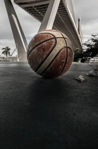 stock image Basketball ball on the Old cement court floor at Outdoor park with Suspension bridge and Sky in the background. Space for text, Selective focus.