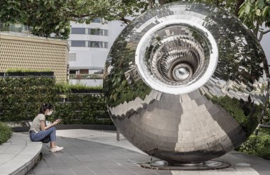 Kimliği belirlenemeyen, kot pantolon ve koruyucu yüz maskesi takan ve akıllı telefon kullanan genç bir kadın şehir parkında paslanmaz çelik öğrenci şeklindeki Spa heykelleriyle oturuyor.