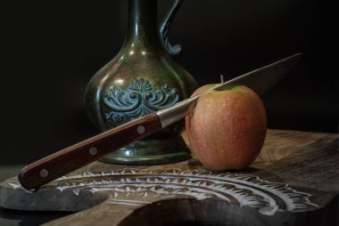 A knife is cutting into Naturally pink fresh kissabel apple fruit and Antique iron jug on wooden cutting board with dark background. Light and Shadow, Space for text, Selective Focus. clipart