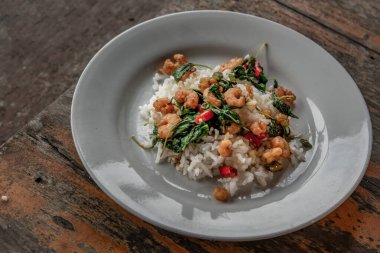 Üzerinde Tayland usulü dünyaca ünlü bir lezzet olan tahta masada servis edilen beyaz tabakta servis edilen Tayland usulü kızarmış fesleğenle karıştırılmış Tayland fesleğeni ve cüce karidesi var..