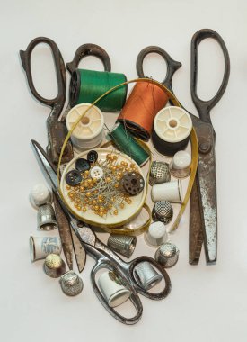 Çelik makas, küçük ahşap sandıkta yüksük, çeşitli renklerde iplik makaraları, yeşil minyatür makaralara iğneler, iğne iplikleri ve mezuralar, beyaz arka planda küçük yuvarlak tepside bulunan raptiyeler. Ögeler dikiliyor