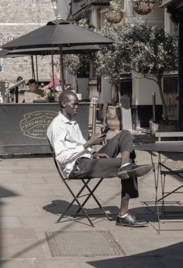 Windsor, İngiltere - Nisan 03, 2025 - Genç bir Afrikalı erkek, beyaz gömlek, siyah pantolon ve deri ayakkabı giyer ve öğleden sonra bir kafede otururken akıllı telefondan mesajları izler. Metin için duvar kağıdı, poster ve boşluk olarak kullan, Selec