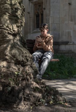 Windsor, İngiltere - Nisan 03, 2025 - Kahverengi kapüşonlu ve kot pantolonlu sarı saçlı bir çocuğun portresi ahşap bir bankta oturuyor ve kilisenin parkındaki akıllı telefonuna gelen mesajlara bakıyor. Metin için Duvar Kağıdı, Poster ve Boşluk olarak kullan Selectiv