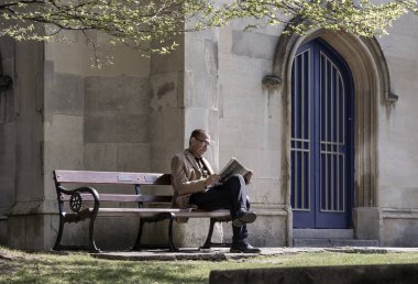 Windsor, İngiltere - Nisan 03, 2025 - Kilise parkındaki ahşap bir bankta otururken gözlüklü yaşlı bir adam gazete okuyor. Emeklilik Zamanı, bunu metin, Seçici odak için duvar kağıdı, poster ve boşluk olarak kullanın.