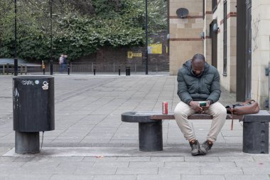 Newcastle, İngiltere - 14 Nisan 2025 - Puffer ceketi giyen kısa saçlı ve kışlık ayakkabılı eşofman altı giyen Afro-Amerikan adam bankta oturuyor ve Tyne üzerine Newcastle 'daki Tyneside' daki akıllı telefonundaki akıllı telefonuna bakıyor. Duvar kağıdın olarak kullan.