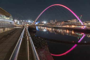 Newcastle, İngiltere - 13 Nisan 2025 - Arka planda Tyne köprüsü ve karanlık gökyüzü olan Iconic Gateshead Milenyum Köprüsü 'nün Nightscape görüntüsü. Nehirde yansıması olan Tyne Nehri 'ni geçen yaya köprüsünü duvar kağıdı, poster, boşluk olarak kullanın..