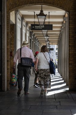 Londra, İngiltere - 13 Mayıs 2025 - Londra 'daki Covent Garden' ın tavanından sarkan simetrik kemerler ve eski moda sokak lambaları arasında el ele yürüyen yaşlı bir çiftin dikey görüntüsü, Romantik ve yaşlılar için sağlıklı yaşam tarzı kavramı.