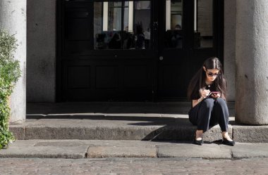 Londra, İngiltere - 13 Mayıs 2025 - Siyah elbiseli ve güneş gözlüklü uzun saçlı bir kadın yürüyüş yolunun kenarında oturuyor ve akıllı telefonundaki görüntülü mesajlara bakıyor. Metin, Seçici odak için Duvar Kağıdı, Poster ve Boşluk olarak kullan.