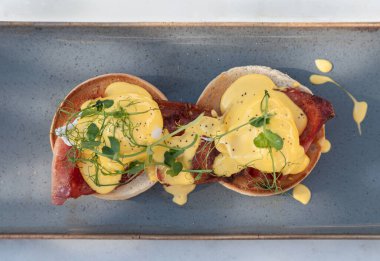 İngiliz çöreği, ızgara domuz pastırması, haşlanmış tavuk yumurtası ve seramik tepside servis edilen holandez sosu ve su teresi ile yapılan yumurtaların üst görüntüsü. Metin, Seçici odak için Duvar Kağıdı, Poster ve Boşluk olarak kullan.