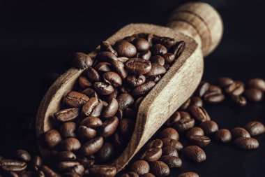 Siyah arka planda ahşap kahve kepçesinde zengin dokulu bir yüzey üzerinde kavrulmuş kahve çekirdekleri manzarası. Yaşamak için sabah kahvenizin tadını çıkarın, onu kopyalama alanınız, tasarım, duvar kağıdı ve poster, seçici odak noktanız olarak kullanın..