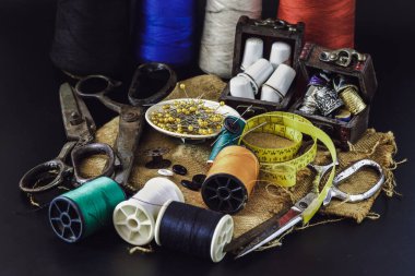 Çelik makas, çeşitli renklerde iplikler, küçük ahşap hazine sandığında yüksük taşları, yeşil minyatür makaralarda sıkışmış iğneler, iplik ve mezura bant, koyu arkaplanlı Çuval Kumaş 'taki küçük yuvarlak tepside iğneler. Ögeler dikiliyor