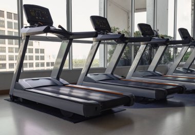 Bangkok, Tayland - 18 Ocak 2025 - Spor salonundaki iki koşu bandının yan görüntüsü. Camdan duvar panellerinden dışarı bakınca manzaralı bina görünüyor. Duvar kağıdı olarak kullan, Poster ve Kopyala alanı, Seçici odak.