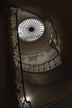 Londra 'daki Somerset Evi' ndeki Nelson Merdiveni 'ne baktığımızda, neoklasik bir binanın içindeki sarmal bir merdiven, üst kısmında belirgin dairesel bir oktulus (tavan penceresi) ve parmaklıklarda karmaşık, süslü bir demiri barındırıyor. Boşluğu kopyala.