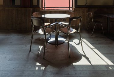 Royal Tunbridge Wells, İngiltere - 29 Nisan 2025 - Güneş ışığı geniş bir pencereden ahşap yemek masalarına, sandalyelere ve boş restoranın arka planında koyu gölgeler bulunan zemine akar. Metin için duvar kağıdı, poster ve boşluk olarak kullan.